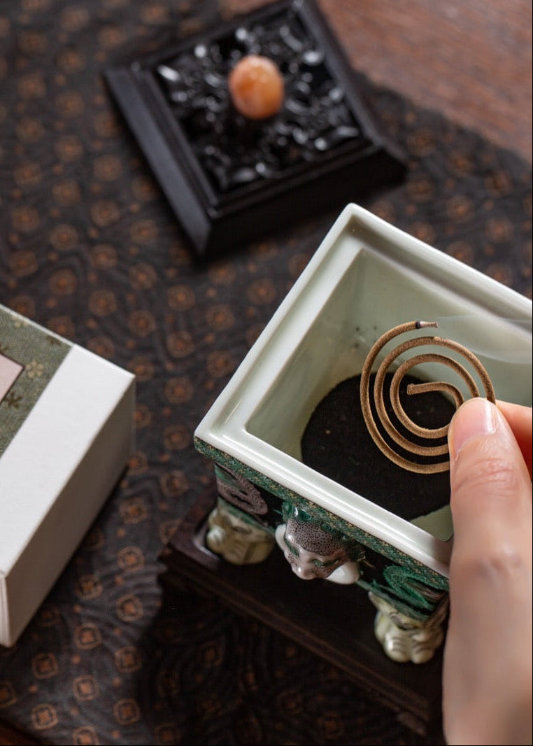 A person's hands are shown holding a white and green box next to an open square ceramic incense burner containing a coiled incense stick, set on a dark patterned table.